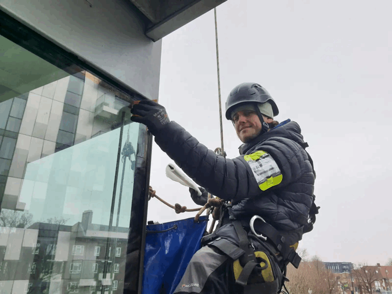 Rope access technician in helmet and harness inspects a glass cladding panel on a high-rise facade.