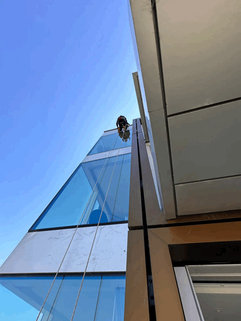 Rope access technician inspects glass and metal cladding facade high on a modern building against blue sky