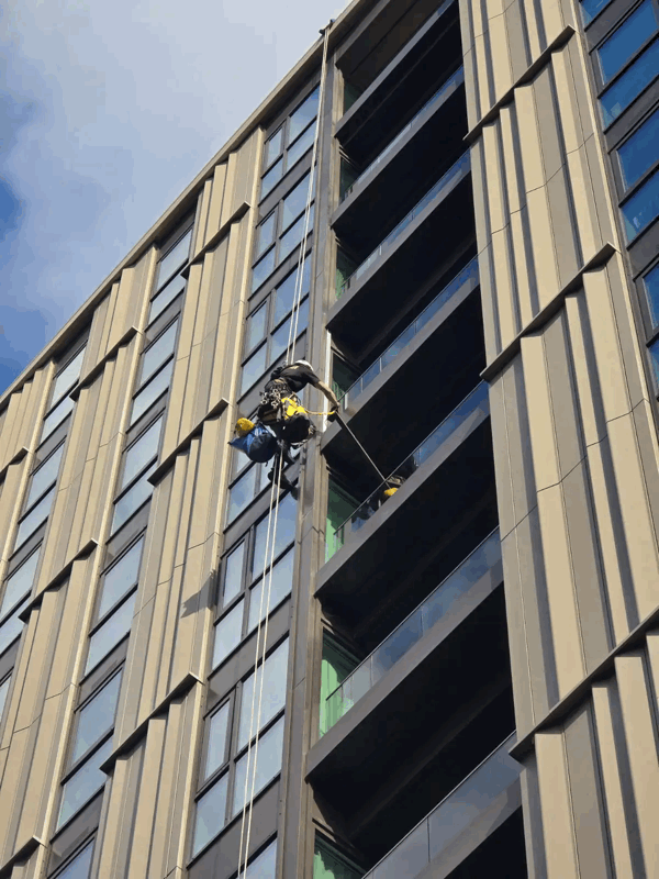 Rope access technician inspects metal cladding panels on a high-rise building facade against a blue sky