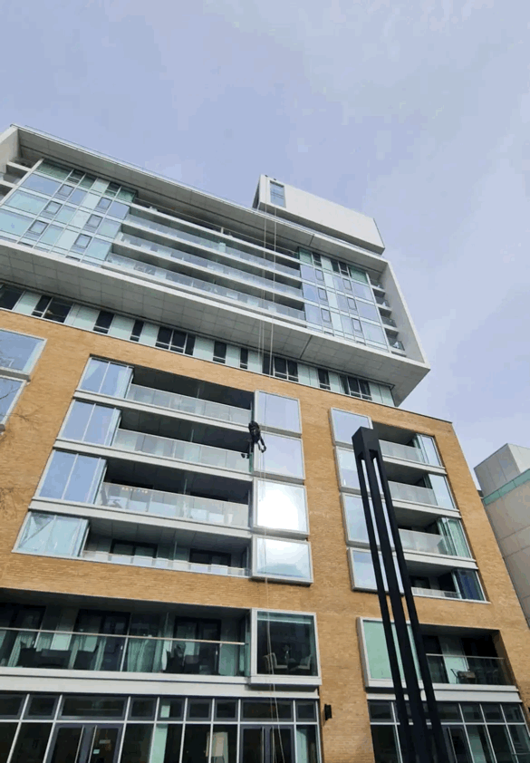 Rope Access Cladding Inspection on High-Rise Building Rope access technician abseiling down a high-rise building to inspect brick-clad and glazed facade panels against a grey sky