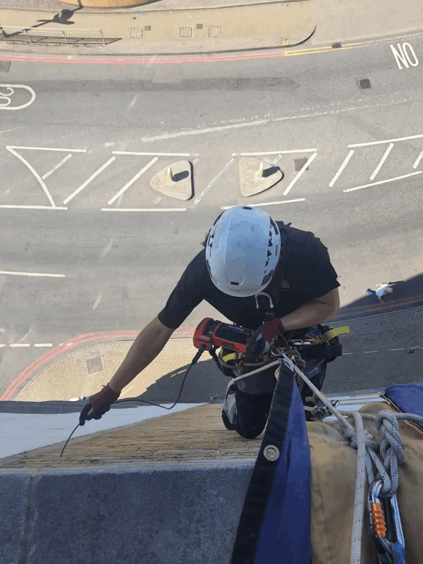 Rope access technician in helmet inspecting building cladding high above a street with markings below