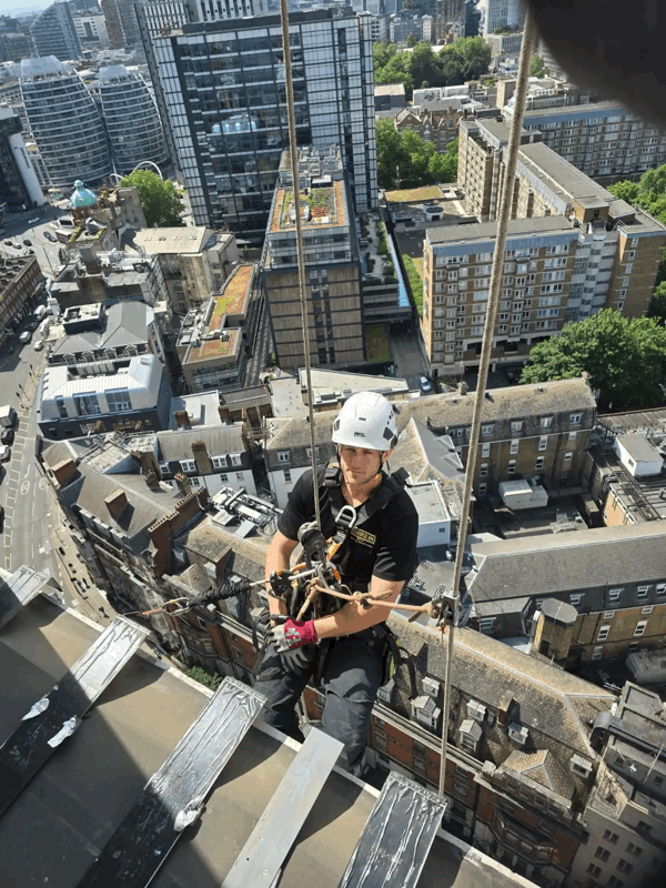 Rope access technician in helmet abseiling down a high-rise building to inspect cladding above the London skyline