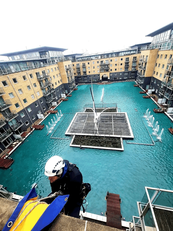 Rope access technician in helmet inspecting building facade above a courtyard with turquoise fountain pool and apartment