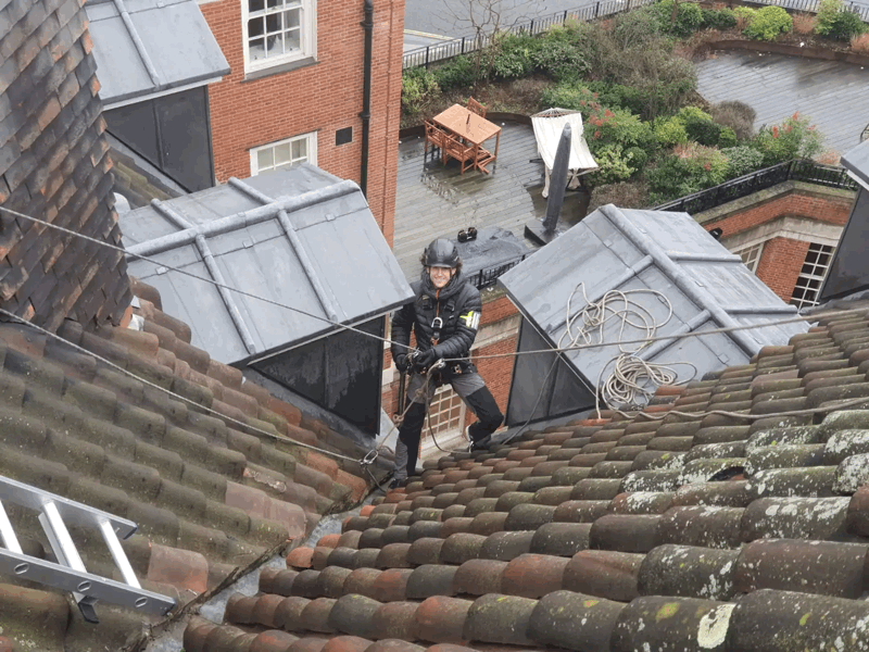 Rope access technician in helmet and harness inspects tiled roof and grey dormer cladding on a brick building