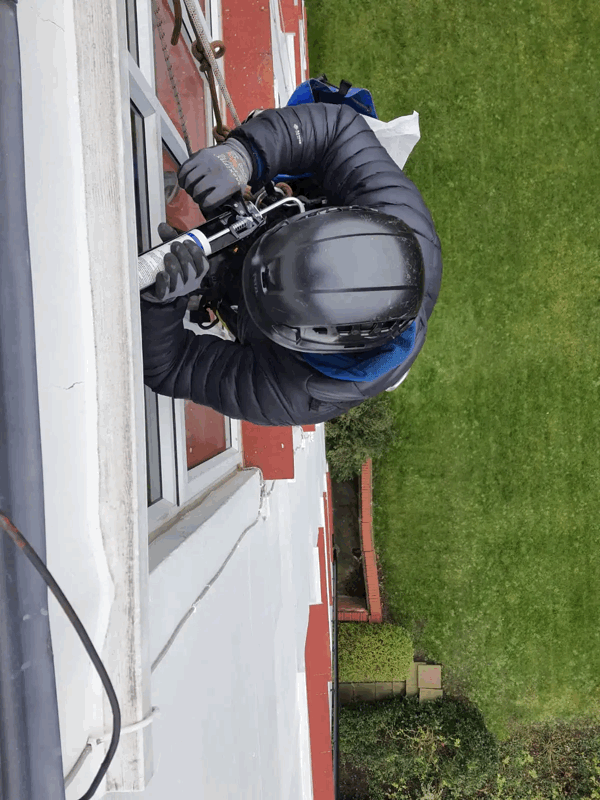 Rope access technician in helmet and harness applies sealant to a window on a red brick building exterior.