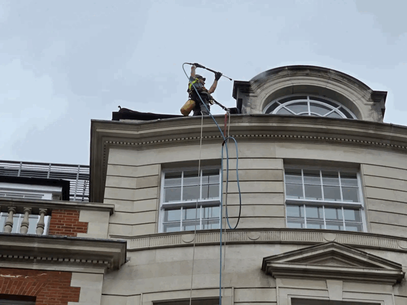 Rope access technician pressure washing a stone building facade near a rounded window on an upper floor