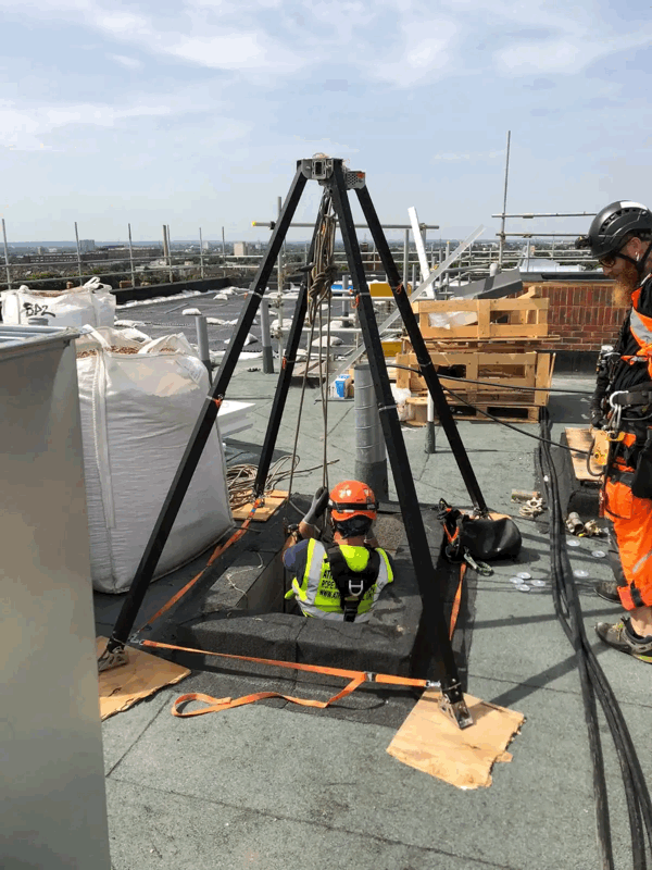Rope access technician descends through a roof hatch using a black tripod rig on a flat commercial rooftop.