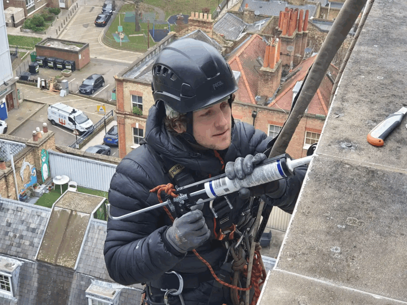 Rope access technician in helmet applies sealant with a caulking gun to a building facade high above a residential street