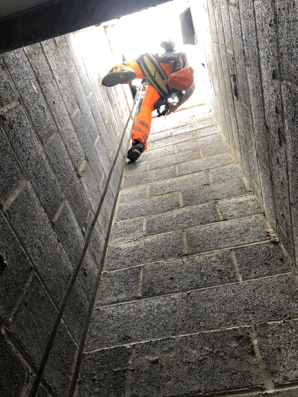 Rope access technician in orange hi-vis descends a narrow concrete cladding shaft during inspection.