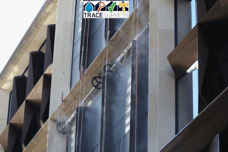 Rope access technicians inspect a high-rise building facade with dark cladding panels and hanging safety ropes.