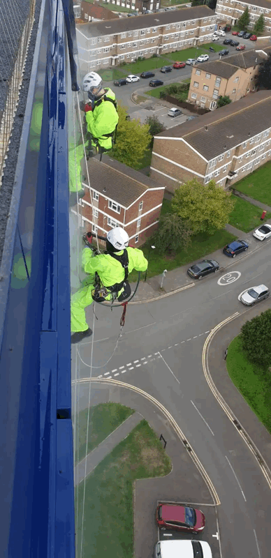 Two rope access technicians in hi-vis suits abseil down a blue-clad high-rise building for cladding inspection above a