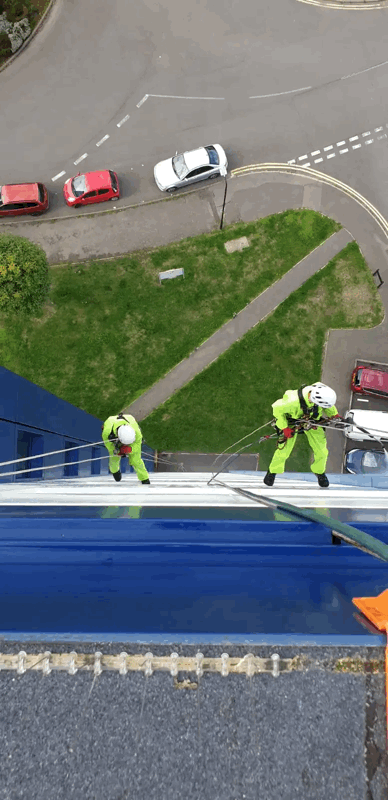 Two rope access technicians in high-vis suits inspect blue metal cladding on a high-rise building exterior.