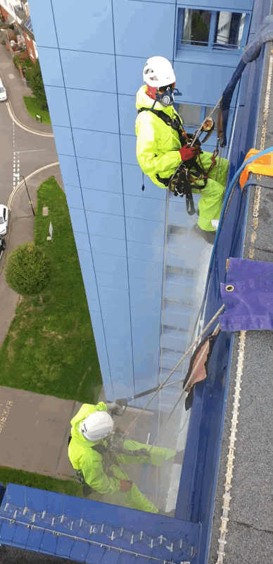 Two rope access technicians in high-visibility suits inspect blue panel cladding on a high-rise building facade