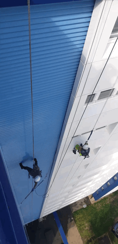 Two rope access technicians inspect blue and white metal cladding on a high-rise building facade.