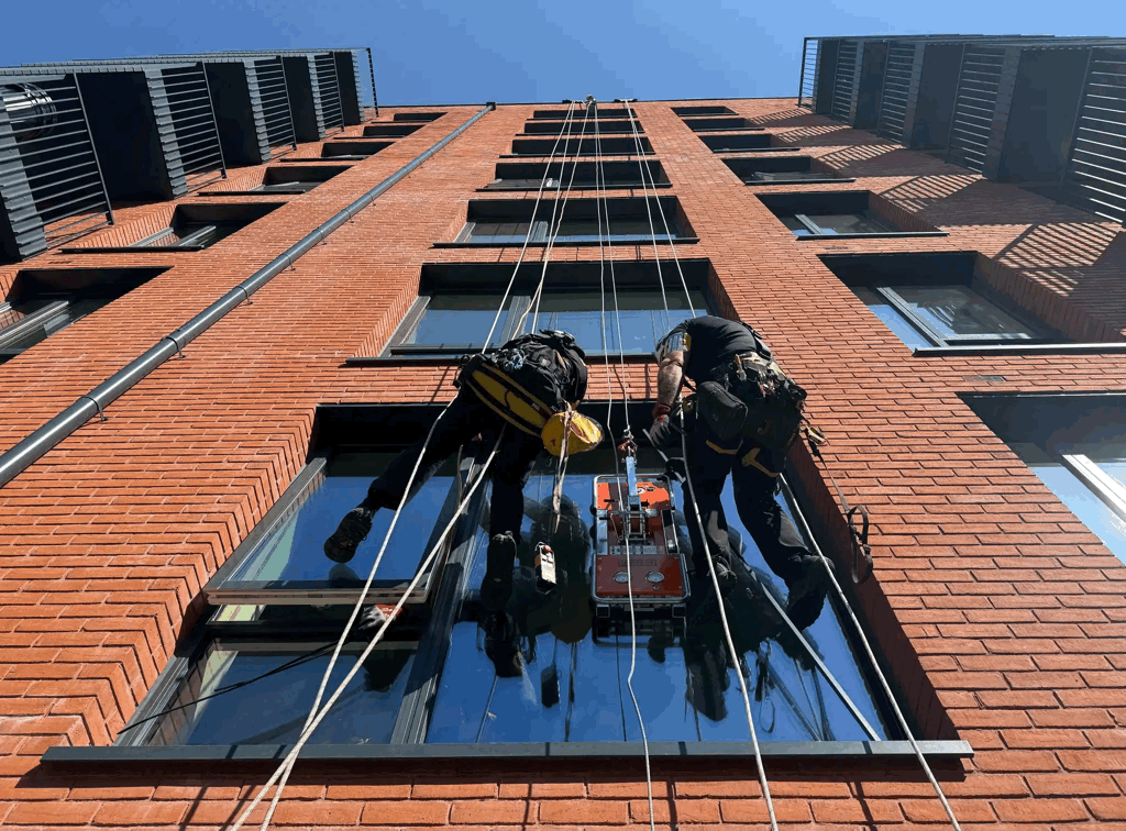Two rope access technicians inspect a red brick-clad high-rise facade window using suspended equipment.