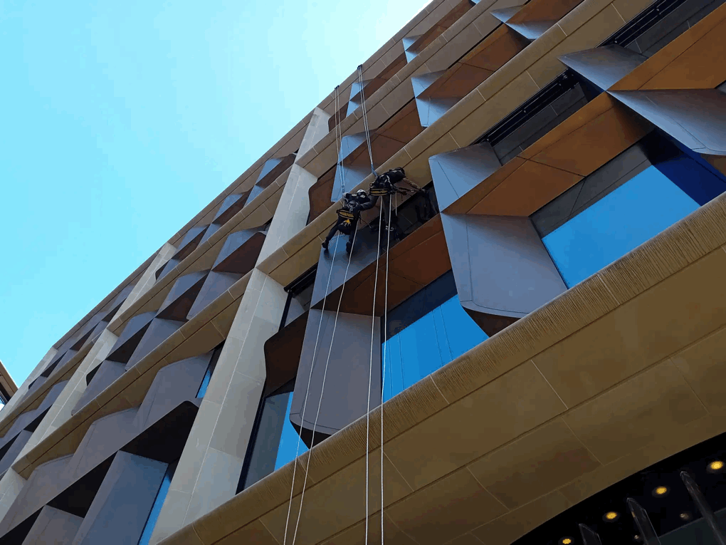 Two rope access technicians inspect tan and grey cladding panels on a high-rise building facade.