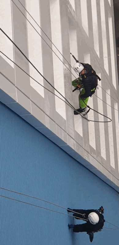 Two rope access technicians in helmets abseil down a striped facade to inspect blue metal cladding panels.