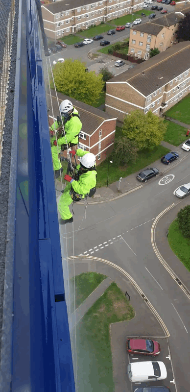 Two rope access technicians in high-vis suits inspect blue cladding panels on a high-rise building above a residential