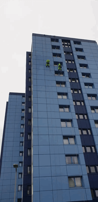 Two rope access technicians in high-visibility gear inspect blue panel cladding on a tall residential tower.
