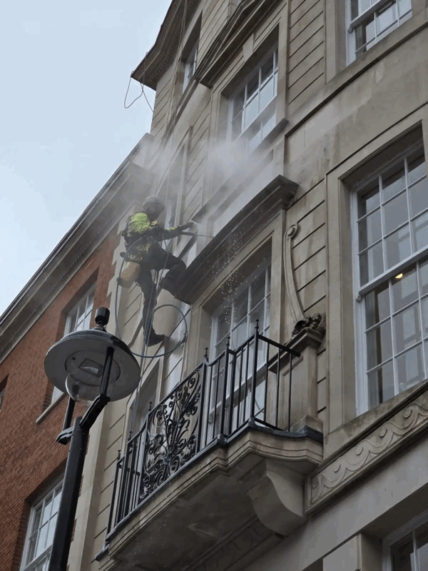 Rope access technician in harness power washes a stone building facade above a wrought iron balcony.