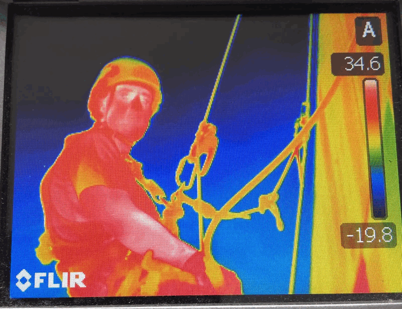 Thermal imaging camera screen shows a rope access technician inspecting building cladding fixings for heat variation